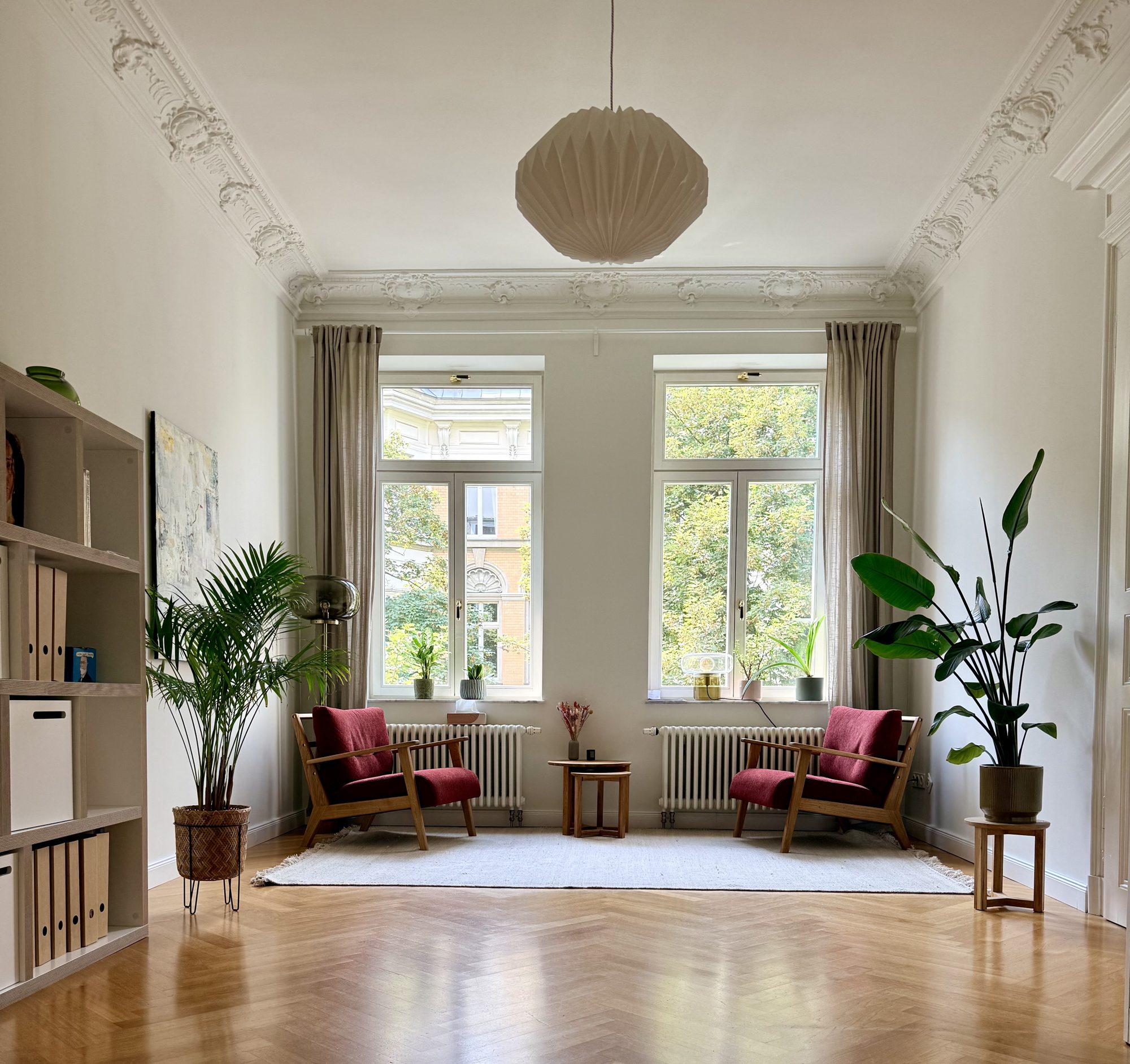 Heller Altbau-Therapieraum mit hoher Stuckdecke, zwei roten Sesseln vor zwei großen Fenstern und Pflanzen.
